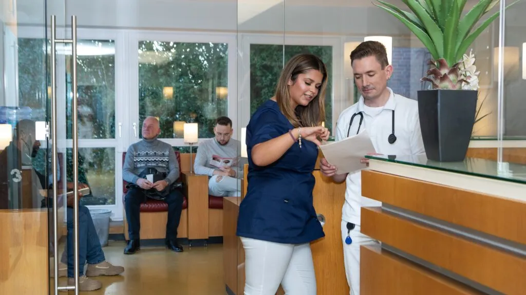 Zwei medizinische Fachkräfte besprechen Unterlagen an einem Empfangstresen, während zwei Patienten im Wartebereich sitzen.
