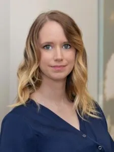 Eine Person mit blonden, welligen Haaren und einem freundlichen Lächeln, die ein dunkles Oberteil trägt.