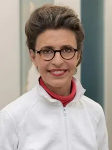 Eine lächelnde Person mit Brille und kurz geschnittenen Haaren, die ein weißes Jackett und ein rotes Shirt trägt