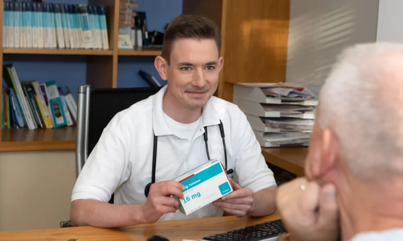 Ein Arzt hält ein Medikamentenpackungsbeispiel mit der Aufschrift 16 mg und unterhält sich mit einer älteren Person.